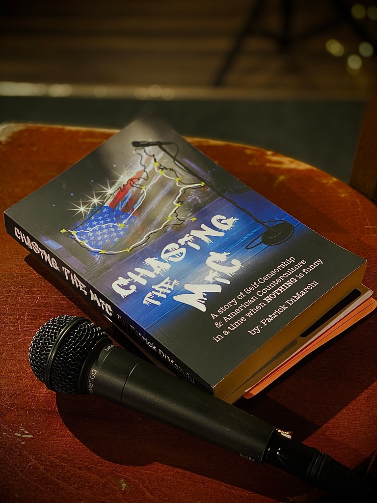 A book sitting on top of a stool next to a microphone at an open-mic comedy night. The title of the book is Chasing The Mic.